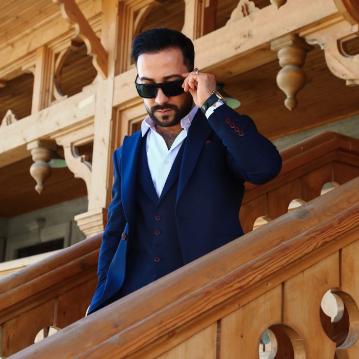 Elegant man in a blue suit and sunglasses descending wooden stairs with ornate architecture.